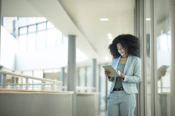 a young female businesswoman stands in the corridor of a modern business interior looking at her digital tablet . The lighting is bright and there is a glow from the tablet device on her face. She is wearing a smart grey business suit. Cloud cost management