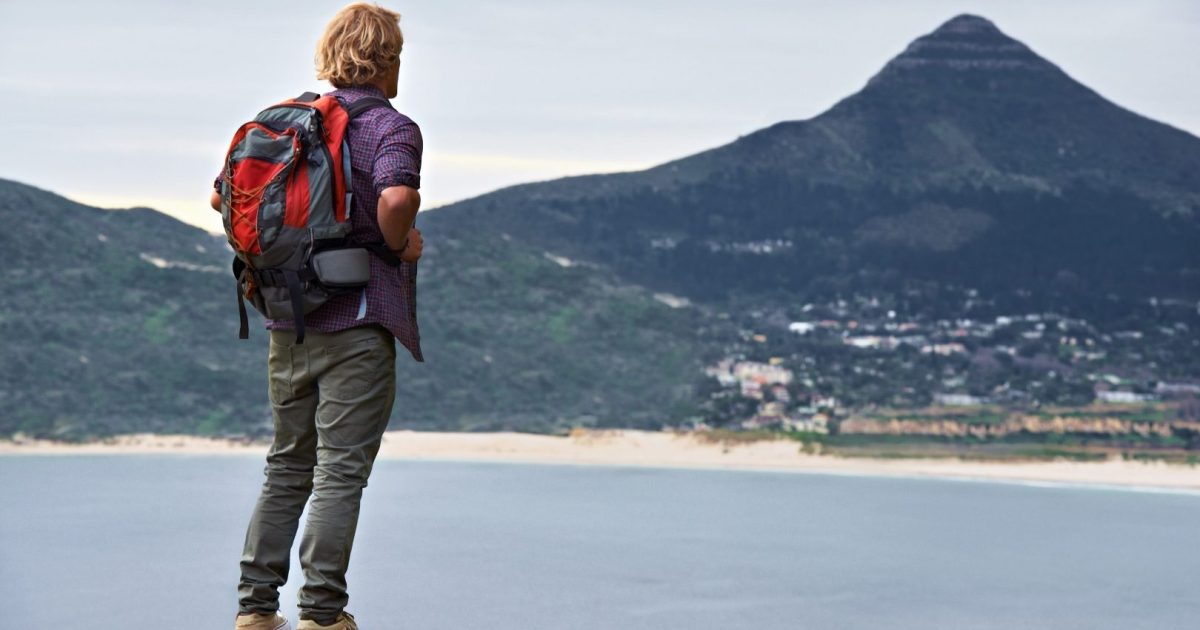 A hiker with a red backpack standing in on a rock overlooking a bay and mountains.